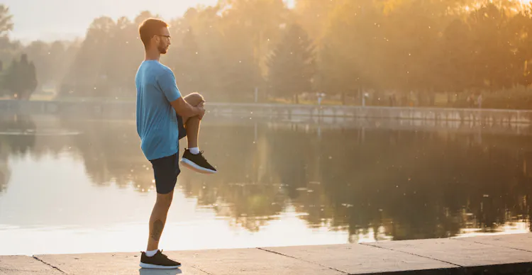 Young man in sportswear stretching by the lake in a city park. R